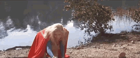 a woman in a red cape is kneeling on the ground near a lake