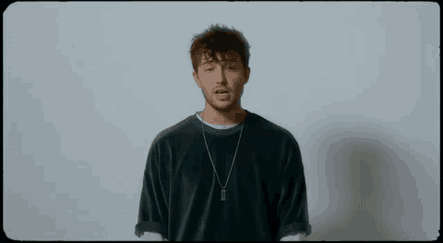 a young man wearing a green sweater and a necklace is standing in front of a white background .