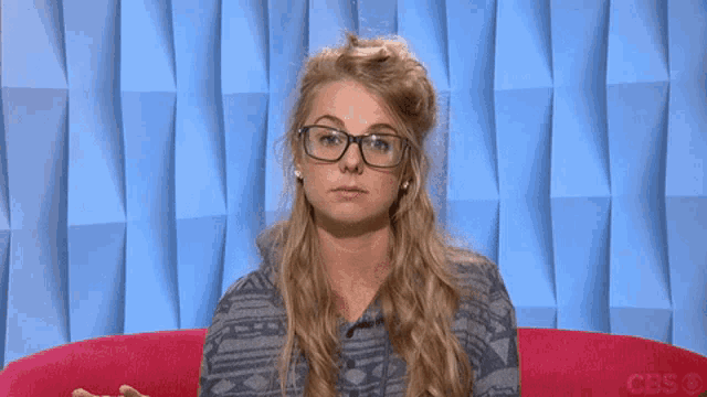 a woman wearing glasses is sitting on a pink couch with a cbs logo in the corner