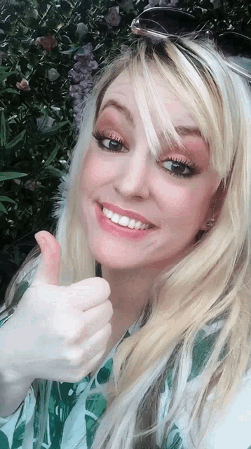 a blonde woman giving a thumbs up in front of some flowers