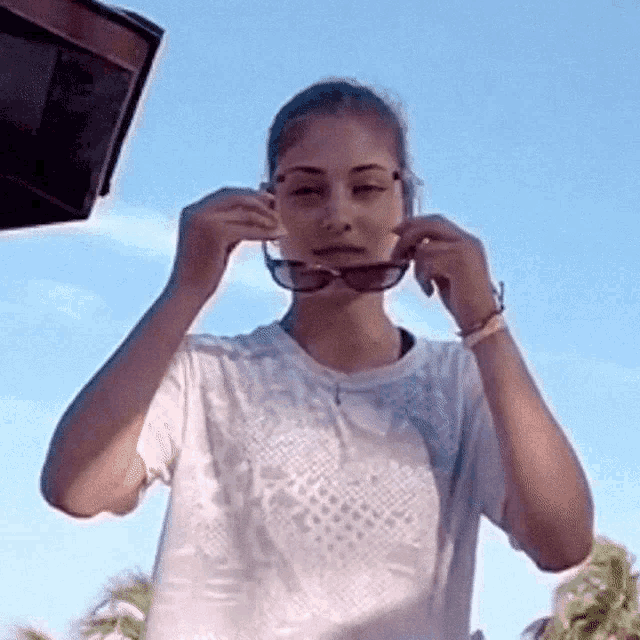 a woman in a white shirt adjusts her sunglasses in front of a blue sky