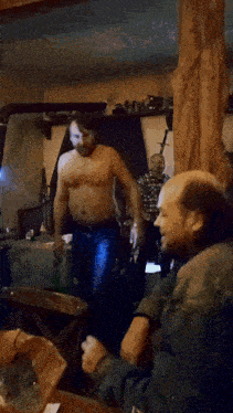 a man without a shirt is standing next to another man who is smoking a cigarette
