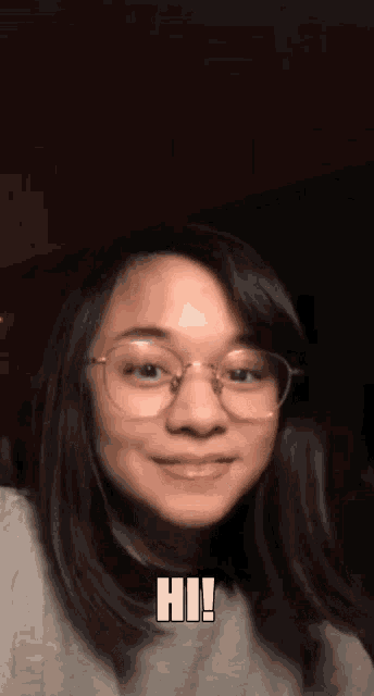 a girl wearing glasses and a gray sweater is smiling and says hi