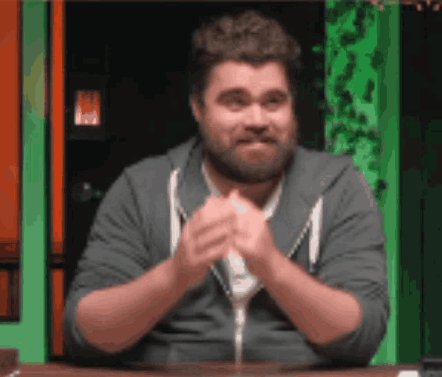 a man with a beard is sitting at a table with his hands folded in front of him