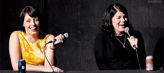 two women are sitting at a table holding microphones and laughing .
