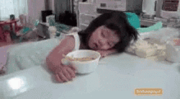 a little girl is sleeping at a table with a bowl of cereal in her hand .