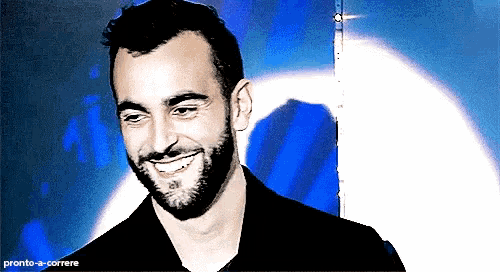 a man with a beard is smiling in front of a blue background with the words pronto-a-correre at the bottom