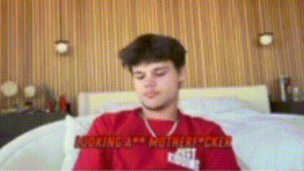 a man in a red shirt is sitting on a bed in a bedroom .