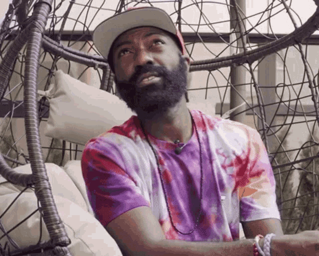 a man with a beard wearing a tie dye shirt sits in a chair