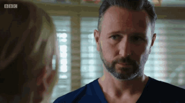 a man with a beard is looking at a woman with the bbc logo visible