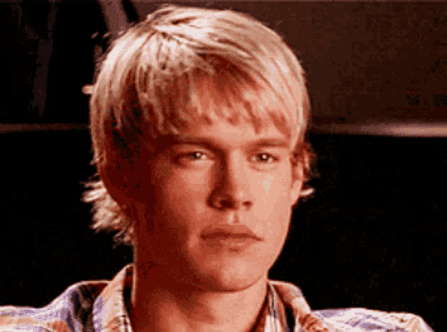 a young man with blonde hair and a plaid shirt looks at the camera