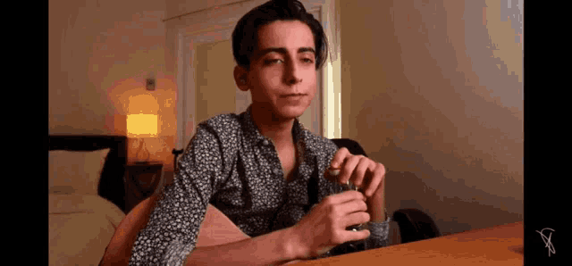 a young man is sitting at a table in a bedroom holding a glass of water .