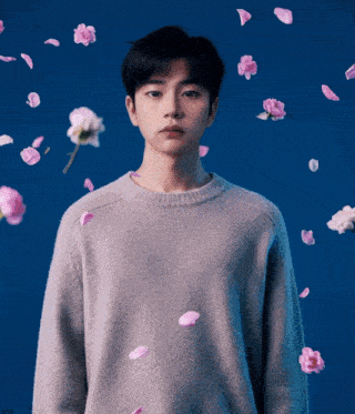 a young man in a grey sweater is surrounded by pink flowers on a blue background