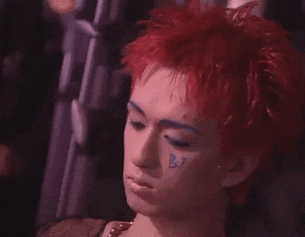 a man with red hair and blue eye makeup is wearing a pearl necklace and a black shirt .
