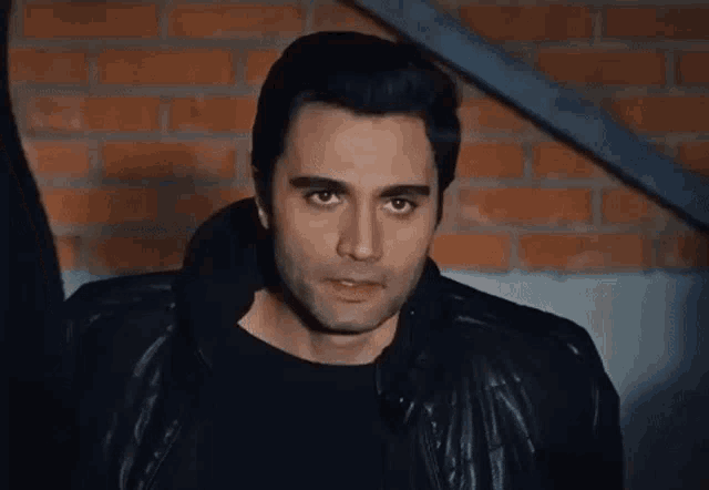a man wearing a black jacket and black shirt stands in front of a brick wall