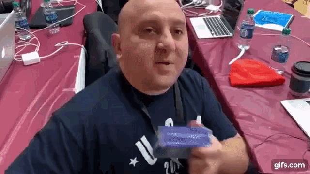 a bald man in a blue shirt is sitting at a table with bottles of water .