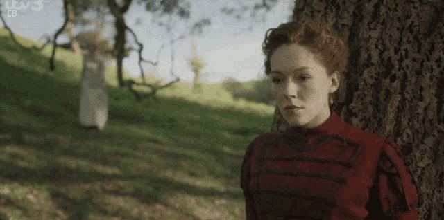 a woman in a red dress leans against a tree in a park