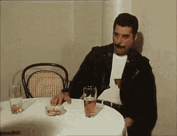 freddie mercury is sitting at a table with a glass of beer and a cigarette .