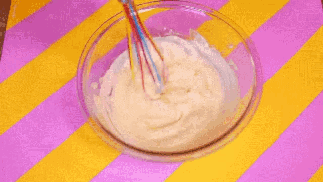 a glass bowl filled with whipped cream and a whisk on a pink and yellow striped surface .