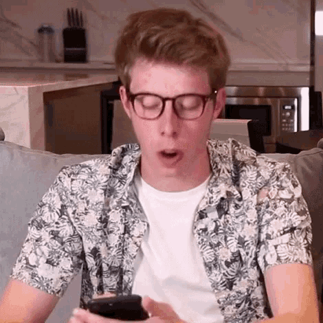 a young man wearing glasses and a floral shirt is sitting on a couch looking at his cell phone .