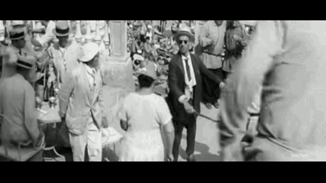 a black and white photo of a man in a suit and tie holding a stick in a crowd of people .