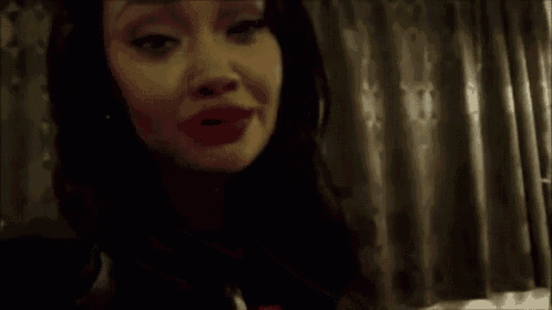 a woman is making a funny face while holding her hand to her chest in front of a curtain .