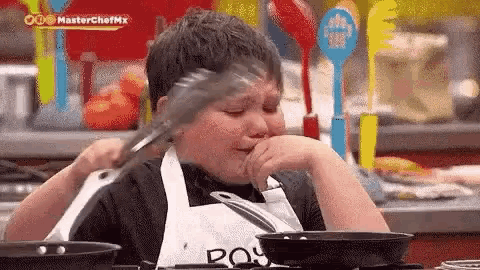a young boy in an apron is holding a tong in his mouth while cooking .