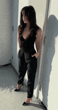 a woman in a black jumpsuit is leaning against a white wall