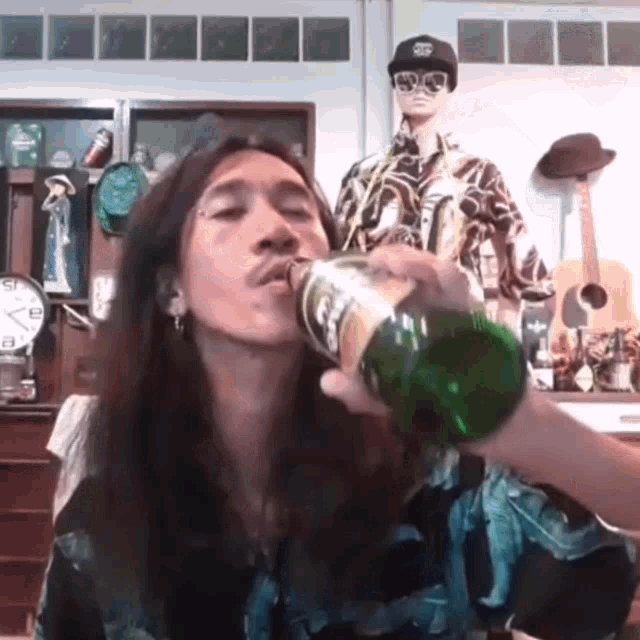 a man with long hair is drinking from a green bottle in front of a clock