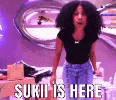 a woman with curly hair is standing in front of a purple wall and says sukii is here .