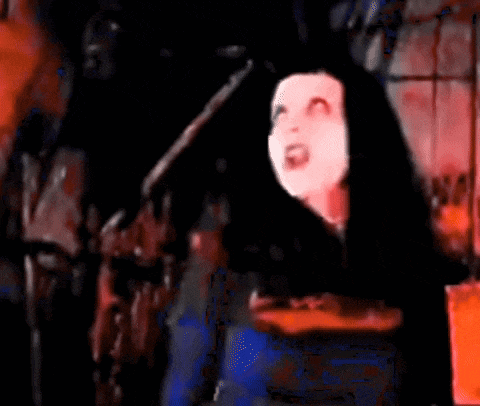 a woman in a white mask is standing in front of a fire in a dark room with a red background .