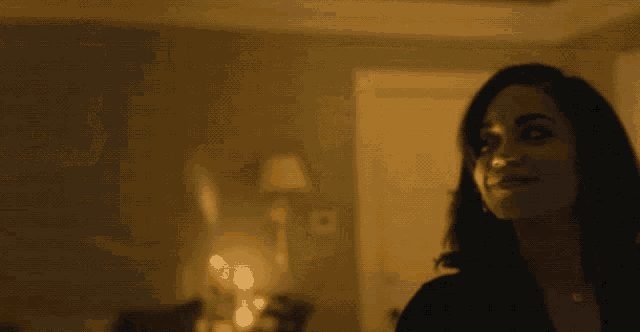 a woman is smiling in a dark room with a lamp behind her