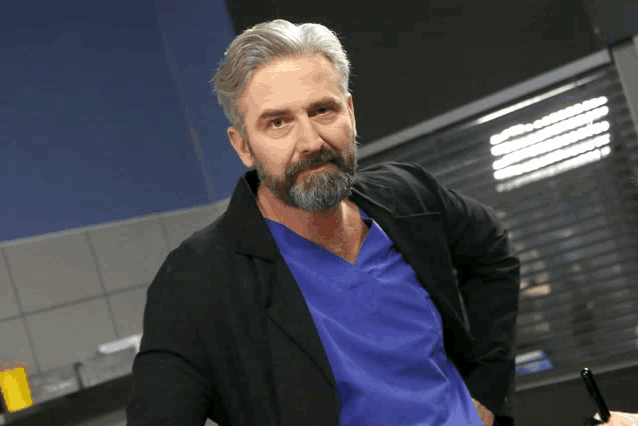 a man with gray hair and a beard is wearing a blue scrub top and black jacket