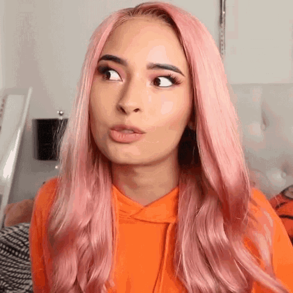a woman with pink hair is wearing an orange sweatshirt and making a funny face .