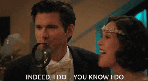 a man in a tuxedo stands next to a woman singing into a microphone and says " indeed i do "