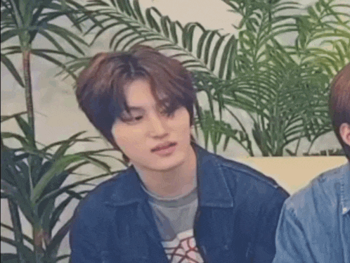 a young man in a denim jacket is sitting next to another young man in front of a palm tree .