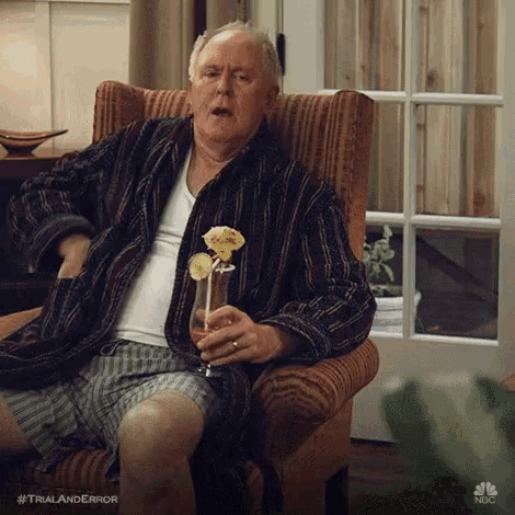 a man in a robe is sitting in a chair with a glass of wine and a flower in his hand .