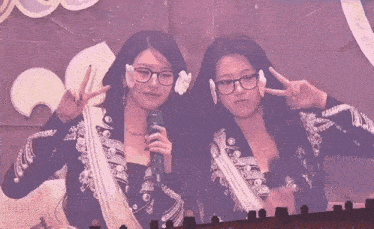 two women wearing glasses and earrings pose for a picture while one holds a microphone