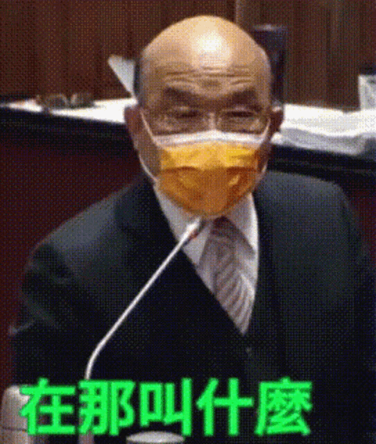 a bald man in a suit and tie is wearing a yellow face mask and speaking into a microphone .
