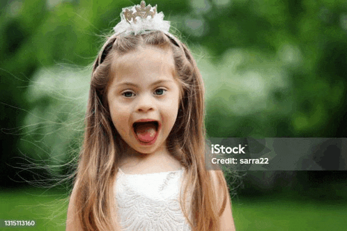 a little girl with down syndrome wearing a tiara and a white dress is making a funny face .