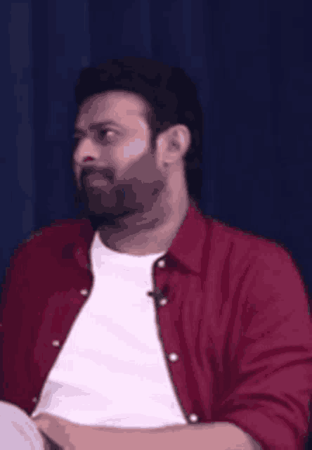 a man with a beard wearing a red shirt and a white shirt is sitting in front of a blue curtain .