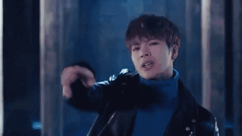 a young man wearing a blue turtleneck and a leather jacket is standing in a dark room .