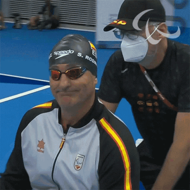 a man wearing a speedo swim cap smiles while another man wearing a mask looks on