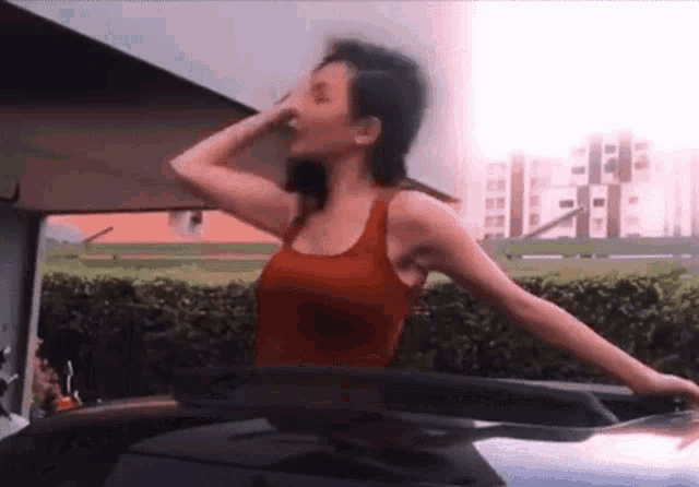 a woman in a red tank top is standing on top of a car with her arms outstretched .
