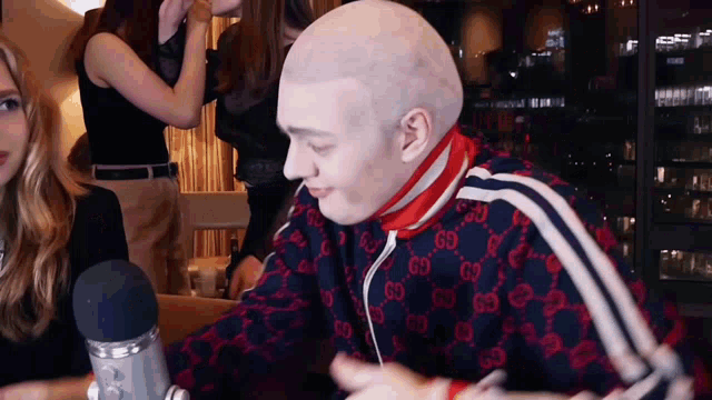a bald man wearing a gucci jacket is sitting in front of a microphone