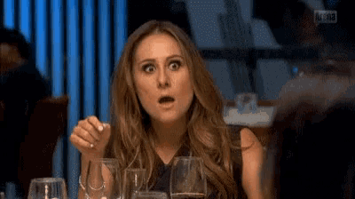 a woman is sitting at a table with glasses of wine and making a funny face .
