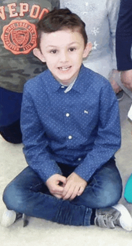 a young boy wearing a blue shirt and jeans is sitting on the floor with his legs crossed .