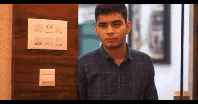 a young man in a plaid shirt is standing in front of a wall with a switch on it