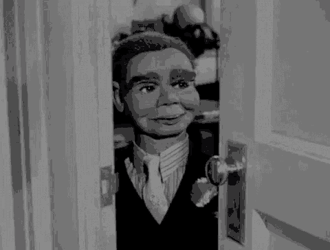 a black and white photo of a puppet in a suit and tie looking out of a doorway .