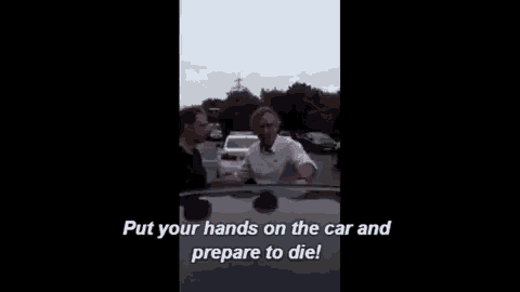 two men are standing next to each other in a parking lot and one of them is telling the other to put their hands on the car .